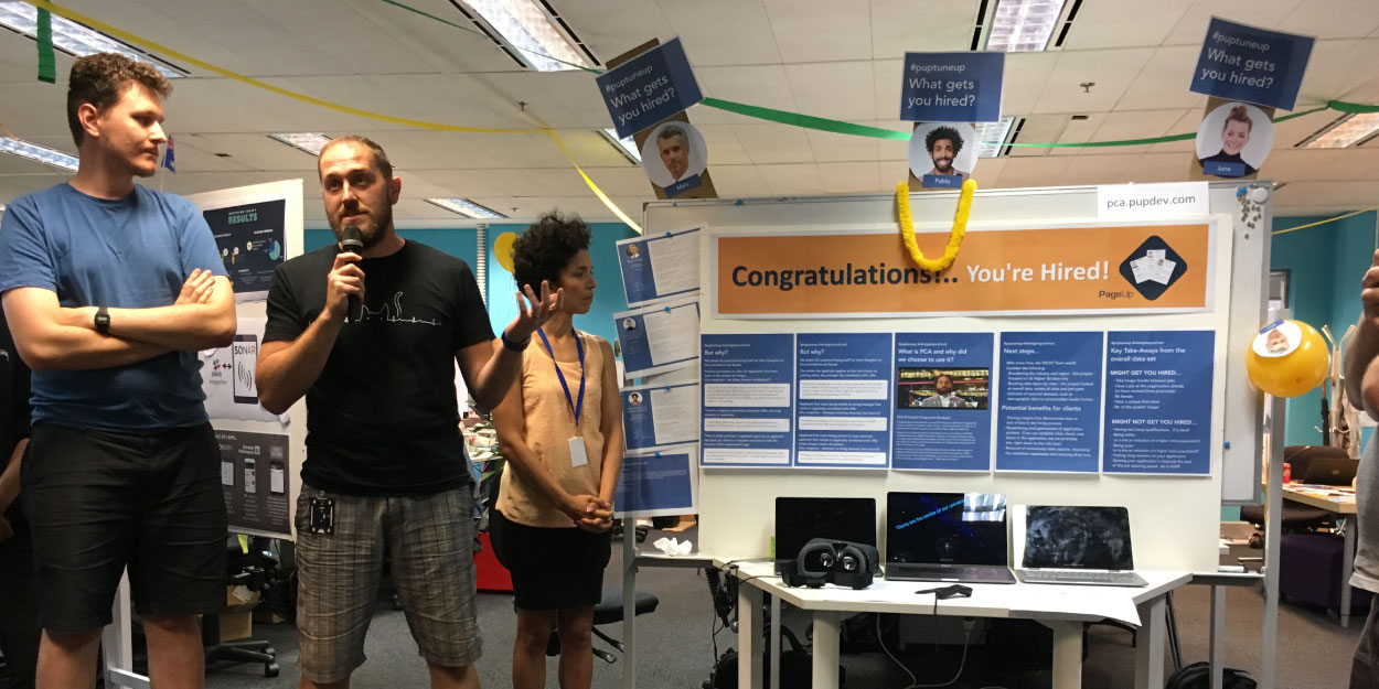 A man with a microphone stands in front of a Hackathon stall. At the stall a woman is looking at a sign that says 'Congratulations, You're Hired!'